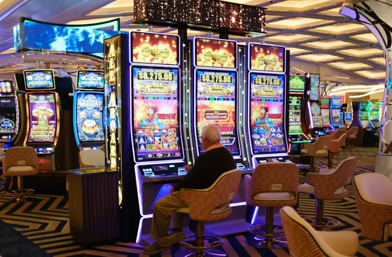 An elderly man sitting at a slot machine in a casino with colorful machines around him